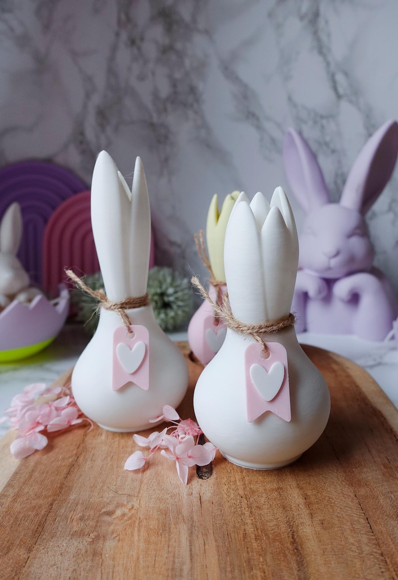K&ouml;nnte beinhalten: Wei&szlig;e Keramikvasen mit tulpenf&ouml;rmigem Design, mit Bindfaden gebunden und mit rosa herzf&ouml;rmigen Anh&auml;ngern verziert. Auf einem Holzbrett, mit einem pastellfarbenen Osterthema.