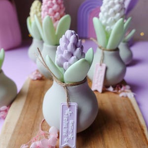 May include: A collection of decorative hyacinth flower sculptures in pastel colors, arranged on a wooden board. Each sculpture features a flower head in shades of pink, purple, and white, with green leaves and a tag that says "Hello Fr&uuml;hling".