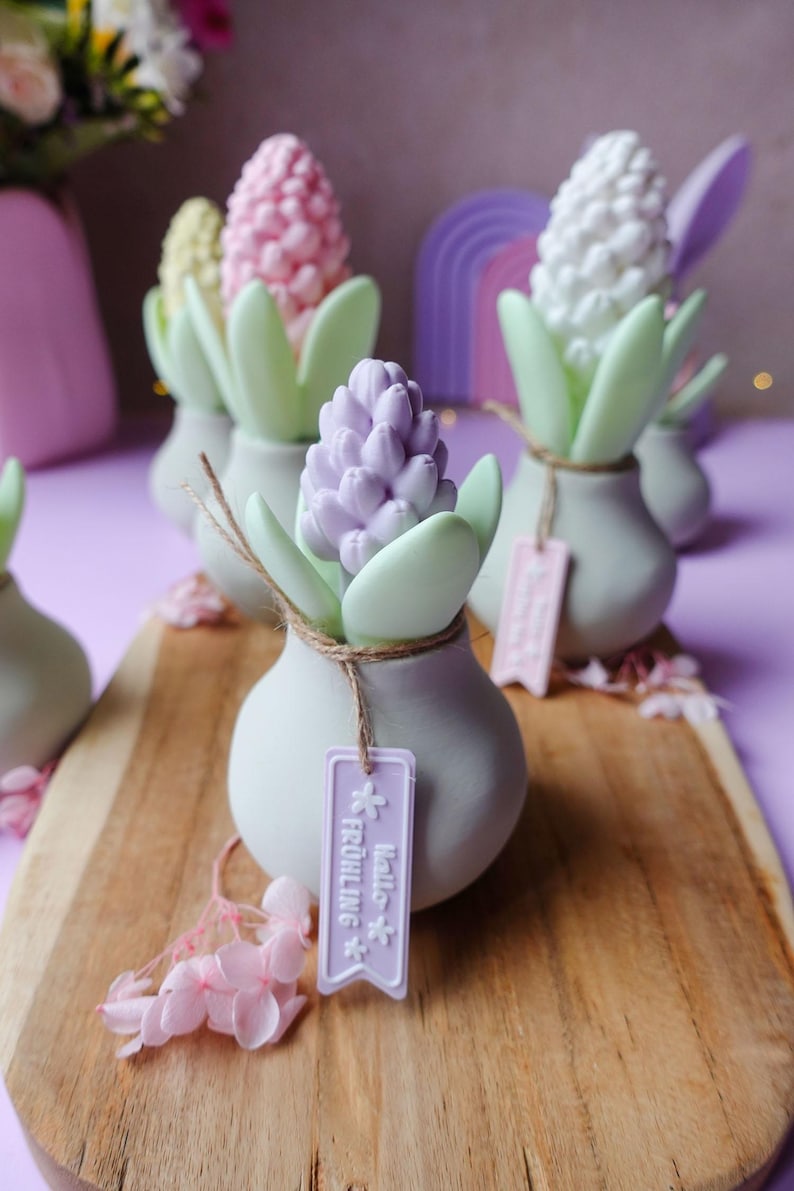 May include: A collection of decorative hyacinth flower sculptures in pastel colors, arranged on a wooden board. Each sculpture features a flower head in shades of pink, purple, and white, with green leaves and a tag that says "Hello Fr&uuml;hling".