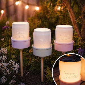 Op de afbeelding: Drie zonne-aangedreven tuinverlichting met witte glazen potkappen. Elke pot staat op een pastelkleurige basis en een houten paal. Eén pot heeft een hart, een andere zegt "Schön HIER" en de derde zegt "Garten ZAUBER".