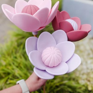 Puede incluir: Tres flores artificiales grandes en tonos rosa y morado. Las flores tienen múltiples pétalos y un capullo central texturizado. Los tallos son marrones y las flores se sostienen contra un fondo de follaje verde.