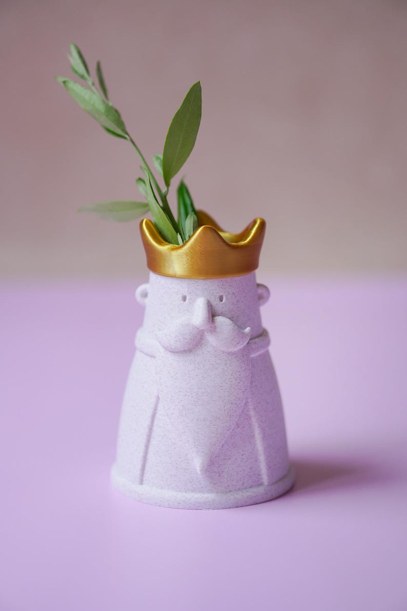 Peut inclure: Un vase mauve clair mouchet&eacute; en forme de personnage avec une moustache et une barbe, surmont&eacute; d'une couronne dor&eacute;e m&eacute;tallique. Le vase pr&eacute;sente de petites oreilles et une barbe pointue, avec un brin de feuilles vertes sortant de l'ouverture.