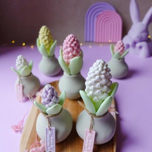 May include: A collection of decorative hyacinth flower sculptures in various pastel colors, including white, pink, yellow, and purple. Each flower sits in a small, light green vase with green leaves. Some vases have small tags attached. The arrangement is on a wooden board.