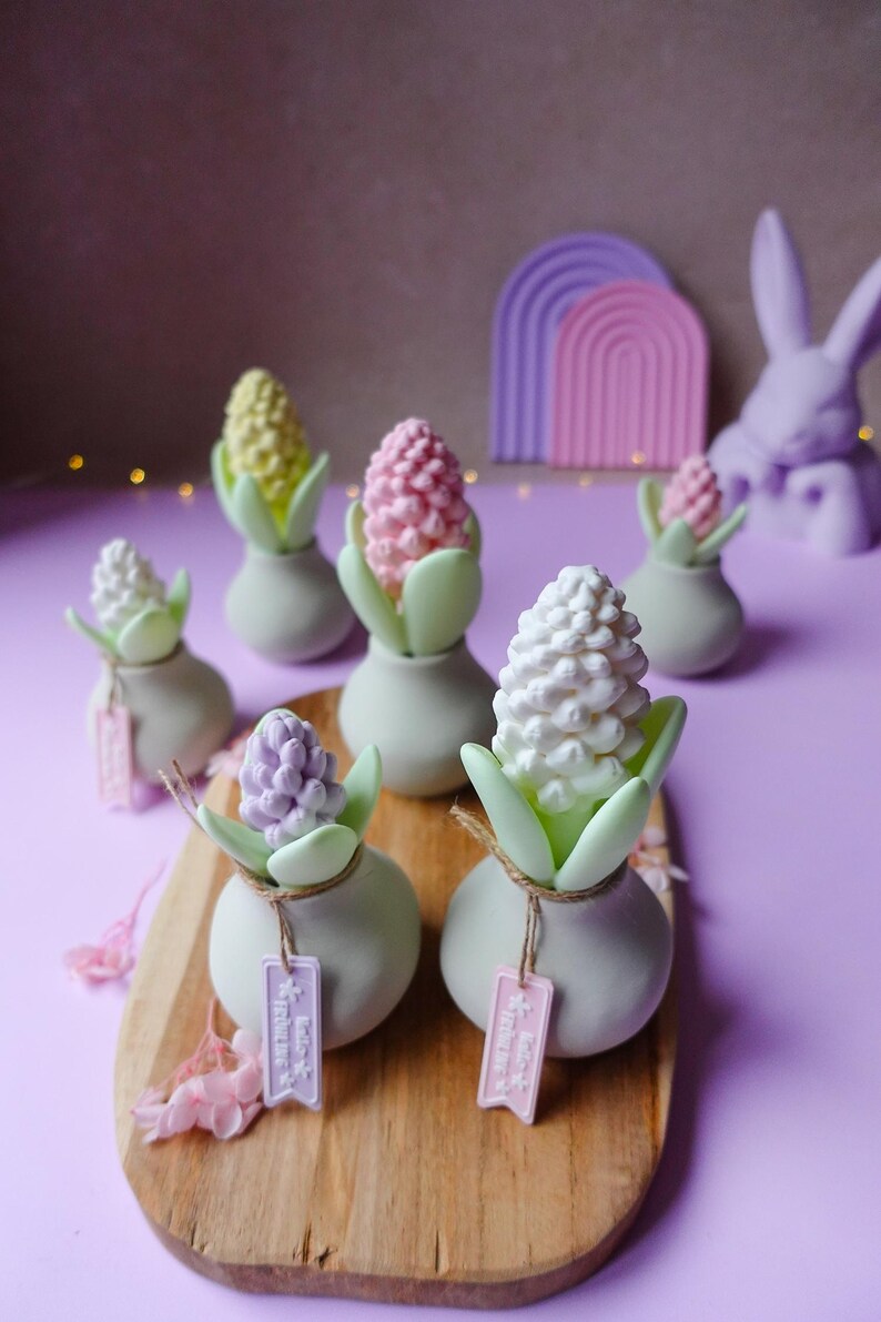 May include: A collection of decorative hyacinth flower sculptures in various pastel colors, including white, pink, yellow, and purple. Each flower sits in a small, light green vase with green leaves. Some vases have small tags attached. The arrangement is on a wooden board.