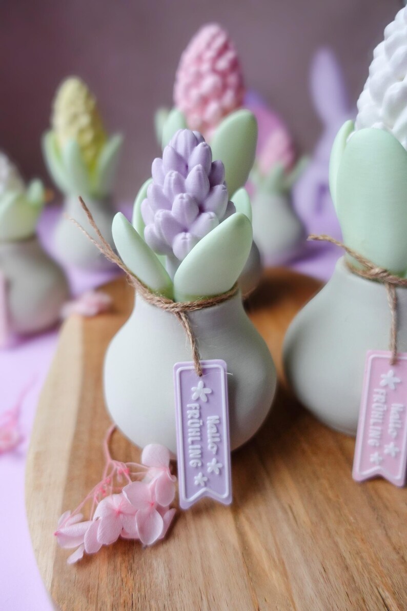 May include: A collection of decorative ceramic vases, each shaped like a bulb, holding colorful hyacinth flowers. The flowers are in shades of purple, pink, yellow, and white, with green leaves. Each vase has a tag with text and is tied with twine.