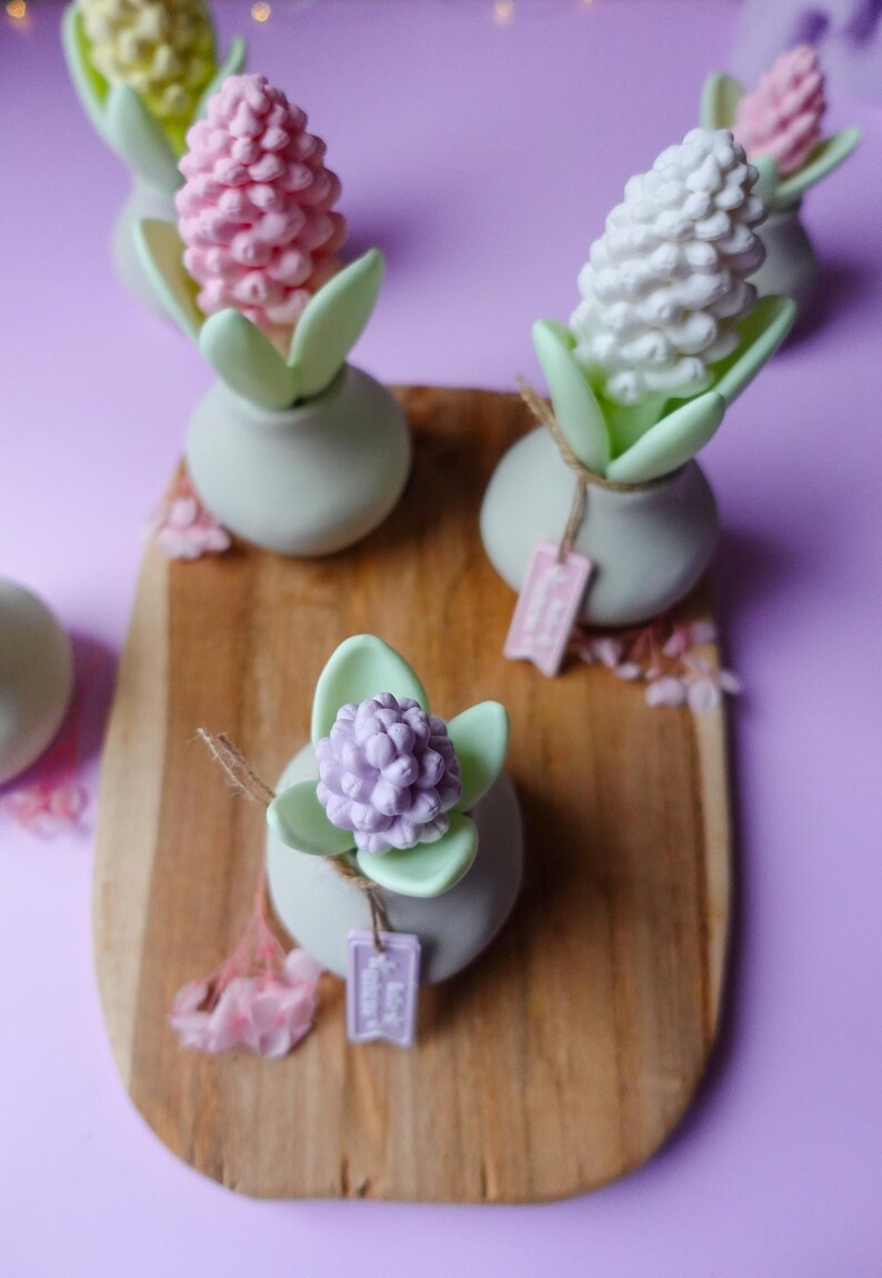 May include: A collection of decorative floral arrangements. Each arrangement features a pastel-colored flower, green leaves, and a small tag, set in a small, light green vase. The arrangements are displayed on a wooden board against a purple background.