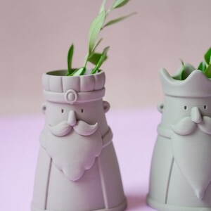 Peut inclure: Deux vases en c&eacute;ramique gris clair en forme de personnages avec moustaches, barbes et coiffes stylis&eacute;es. Chaque vase pr&eacute;sente une base large, une ouverture cylindrique au sommet et contient une petite branche verte feuillue au centre.