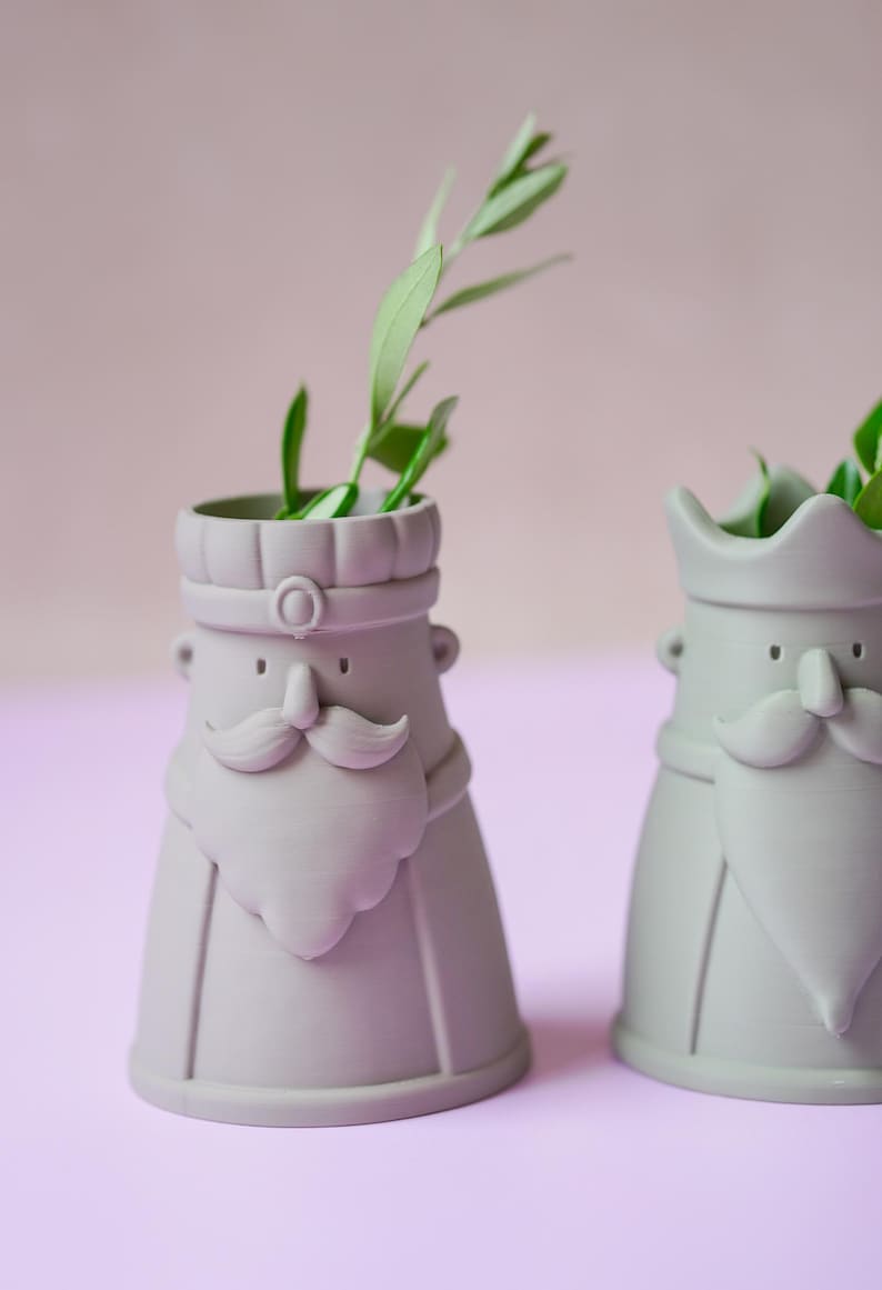 Peut inclure: Deux vases en c&eacute;ramique gris clair en forme de personnages avec moustaches, barbes et coiffes stylis&eacute;es. Chaque vase pr&eacute;sente une base large, une ouverture cylindrique au sommet et contient une petite branche verte feuillue au centre.