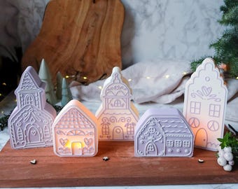 May include: A collection of lavender-colored ceramic house-shaped candle holders. The houses vary in design, with intricate details like windows, doors, and roof patterns. One house glows with a warm light. They are arranged on a wooden board.