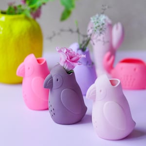 Op de afbeelding: Een verzameling kleurrijke vazen in de vorm van vogels in roze, paarse en gele tinten. Elke vaas bevat een bloem, waardoor een levendige weergave ontstaat. De vazen zijn gerangschikt op een lichtpaars oppervlak, wat een speels en decoratief thema laat zien.