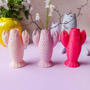 Op de afbeelding: Een verzameling vazen in de vorm van een kreeft in verschillende tinten roze en rood, elk met delicate witte bloemen en takken. De vazen hebben een gedetailleerd ontwerp, met een getextureerd lichaam en klauwachtige armen. Een gele vaas en andere vazen staan op de achtergrond.