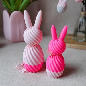 May include: Two decorative Easter bunny figurines. The bunnies are pink and white, with a spiral design. One bunny has a small pink flower. The background includes a pink flower pot with tulips and hanging bunny ornaments.