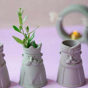 Peut inclure: Trois vases en c&eacute;ramique vert sauge mat en forme de personnages stylis&eacute;s avec moustaches et barbes. Le vase central contient une petite branche feuillue verte. Chaque figurine pr&eacute;sente un couvre-chef distinct, couronne ou turban, et des traits simples.