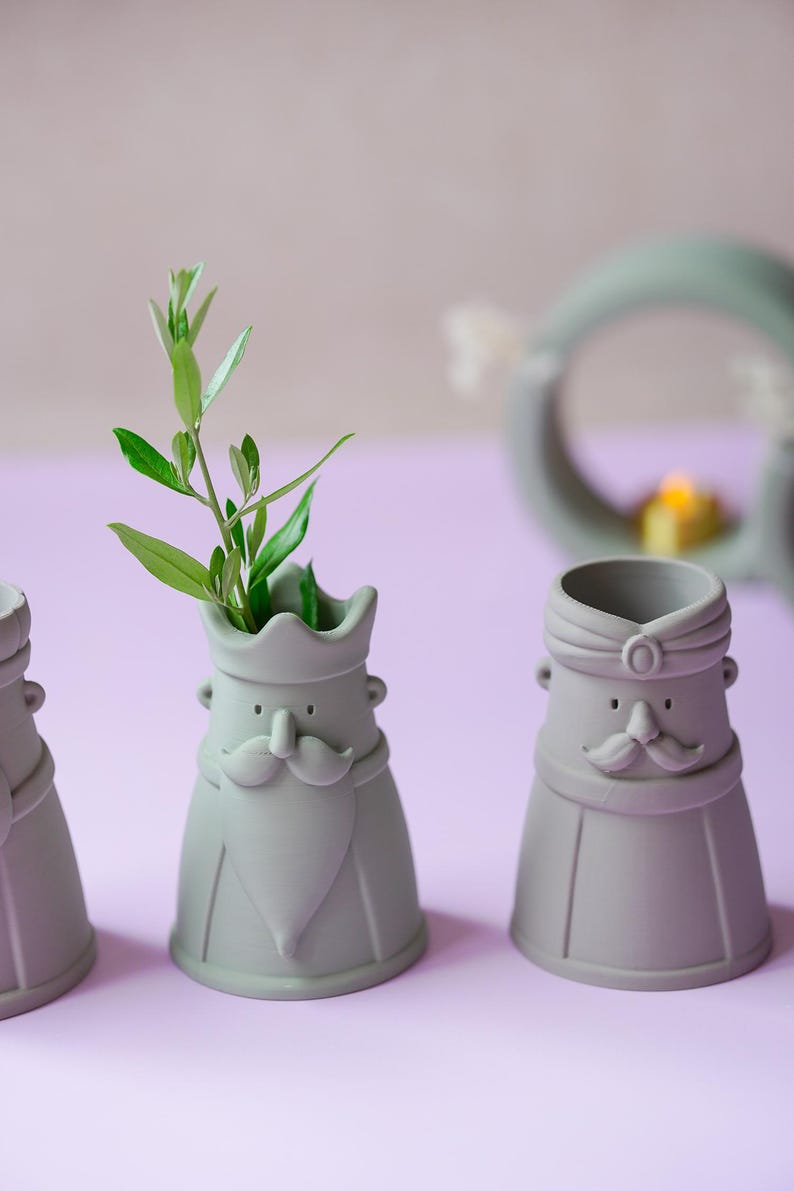 Peut inclure: Trois vases en c&eacute;ramique vert sauge mat en forme de personnages stylis&eacute;s avec moustaches et barbes. Le vase central contient une petite branche feuillue verte. Chaque figurine pr&eacute;sente un couvre-chef distinct, couronne ou turban, et des traits simples.