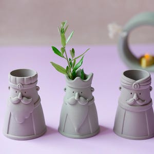 Peut inclure: Trois r&eacute;cipients en c&eacute;ramique gris clair mat en forme de personnages stylis&eacute;s avec moustaches et coiffes. Le r&eacute;cipient central contient un petit brin de feuilles vertes. Chaque figurine a une base large, un corps fusel&eacute; et des traits distincts.
