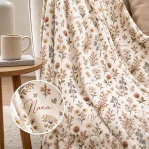 May include: Cream-colored blanket with a floral pattern in shades of brown, yellow, and blue. The word "Nana" is written in a cursive font across the blanket. The blanket is draped over a beige sofa, creating a cozy and inviting aesthetic.