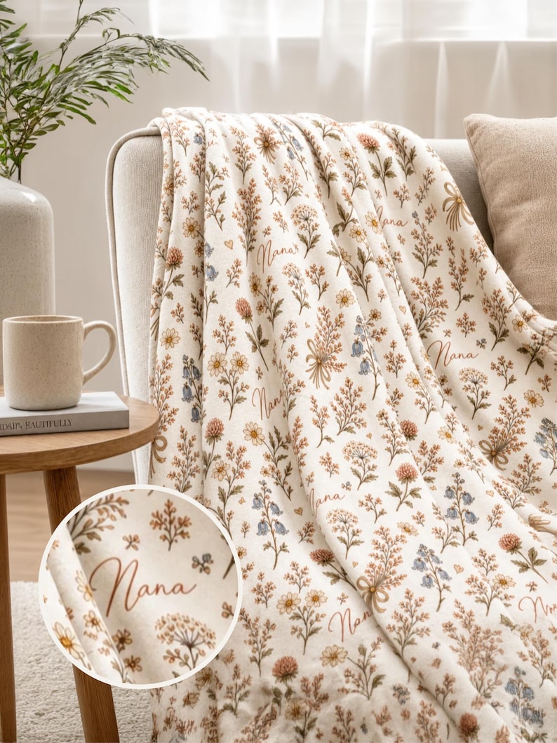 May include: Cream-colored blanket with a floral pattern in shades of brown, yellow, and blue. The word "Nana" is written in a cursive font across the blanket. The blanket is draped over a beige sofa, creating a cozy and inviting aesthetic.