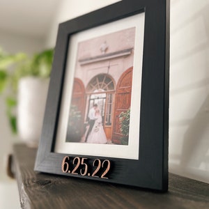 May include: Black picture frame with a wedding photo inside. The frame has a wooden plaque with the date "6.25.22" on the bottom.