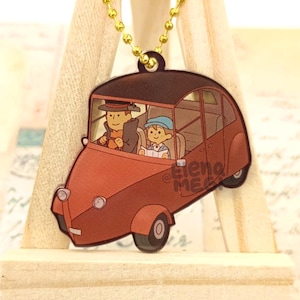 May include: A red and brown cartoon car charm with two figures inside. The car has a white roof and a gold chain attached to the top. The figures are wearing hats and are looking out the windows. The charm is on a wooden surface.