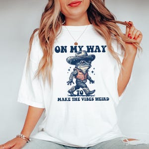 May include: White t-shirt with a graphic of a frog wearing a cowboy hat and the text "On My Way To Make The Vibes Weird".