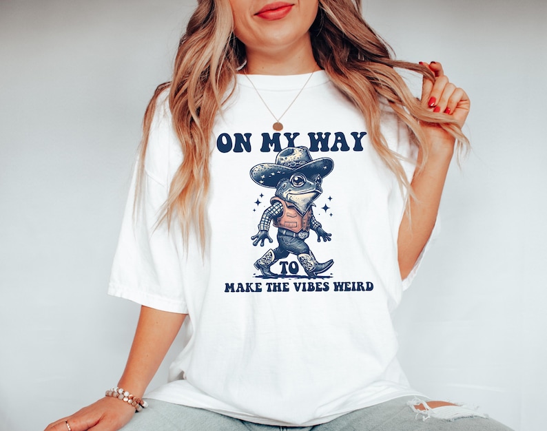 May include: White t-shirt with a graphic of a frog wearing a cowboy hat and the text "On My Way To Make The Vibes Weird".