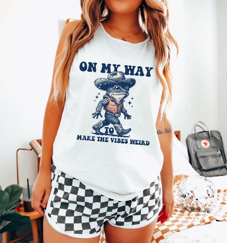 May include: White tank top with a cartoon frog wearing a cowboy hat and vest. The text on the shirt reads "On my way to make the vibes weird".