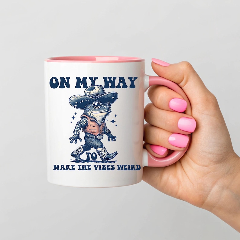 May include: White ceramic mug with a pink rim and handle. The mug features a cartoon frog wearing a cowboy hat and vest, with the text "On my way to make the vibes weird".