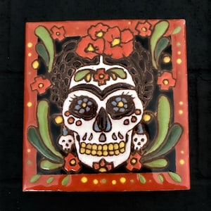 May include: A square ceramic tile featuring a central white skull with black, yellow, and blue decorative details. The skull is surrounded by red flowers, green leaves, and smaller skull accents against a black background, all framed by a red border.