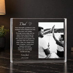 May include: A clear acrylic photo frame with a black and white photo of a father and son giving a high five. The frame has the word "Dad" and a heartfelt message engraved on it.