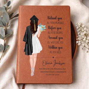 May include: A brown leather-bound journal with a graduation theme. The cover features an illustration of a graduate in a cap and gown, holding flowers. Text includes the graduate's name, degree, university, and class year.