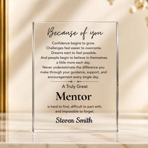 May include: A clear acrylic block with the text "Because of you" and a message about mentorship. The word "Mentor" is prominently displayed, followed by a personalized name. The block is on a reflective surface.