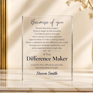 May include: A rectangular clear acrylic plaque featuring black printed text that reads: Because of you, People feel encouraged. Dreams begin to feel possible. Confidence grows stronger. And ordinary moments become opportunities to make a lasting impact. Never underestimate the difference you make through your kindness, leadership, and encouragement every single day. A True Difference Maker is hard to find, difficult to part with, and impossible to forget. Steven Smith.