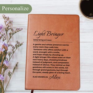 Puede incluir: Un diario de cuero marrón con las palabras "Light Bringer" y una definición impresa en la portada. El diario incluye el nombre "Alex" y está personalizado. Un bolígrafo y una taza de café son visibles en el fondo.