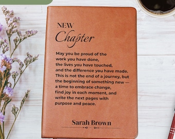 May include: A brown leather journal with the words "New Chapter" and a personalized message. The journal is open, revealing lined pages. A pen and a cup of coffee are on the table.
