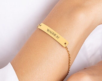 May include: Gold-colored bracelet with the word "WARRIOR" engraved on a rectangular bar. The bar is connected to a delicate chain. The bracelet is shown on a person's wrist, against a white shirt.