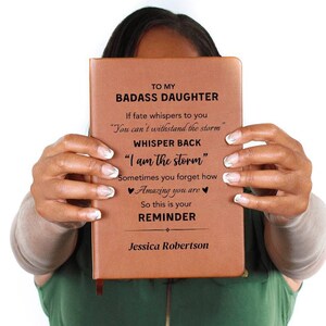以下が含まれることがあります： 「To My Badass Daughter」というテキストと、課題克服に関するインスピレーションを与える引用が書かれた、茶色の革製のジャーナル。ジャーナルは「Jessica Robertson」という名前でパーソナライズされています。