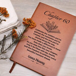 Puede incluir: Un diario de cuero marrón con el texto "Capítulo 60" y un mensaje personalizado. El diario presenta una ilustración de un libro abierto con detalles florales y un marcapáginas de cinta. El nombre "Anna Huang" también está impreso en el diario.