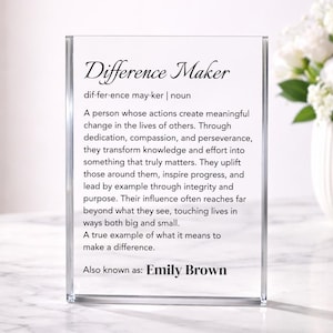 Puede incluir: Un bloque acrílico transparente con las palabras "Difference Maker" en una elegante escritura. La definición de "Difference Maker" está impresa debajo, junto con el nombre "Emily Brown". Un regalo o accesorio de escritorio reflexivo.