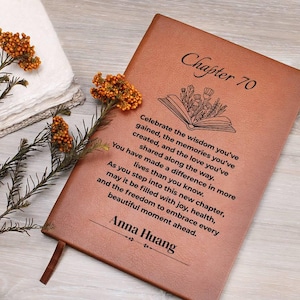 May include: A brown leather journal with the text "Chapter 70" and a personalized message. The journal has a ribbon bookmark and is decorated with dried orange flowers and greenery.