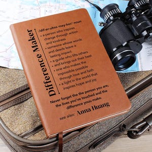 May include: A brown leather journal with the words "Difference Maker" and a definition printed on the cover. The journal includes a ribbon bookmark and is next to binoculars and a map.