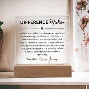 Può includere: Una placca in acrilico trasparente con una base in legno, che presenta il testo "DIFFERENCE Maker" e una citazione sul fare la differenza attraverso piccole azioni. La citazione è attribuita a "Olivia James".