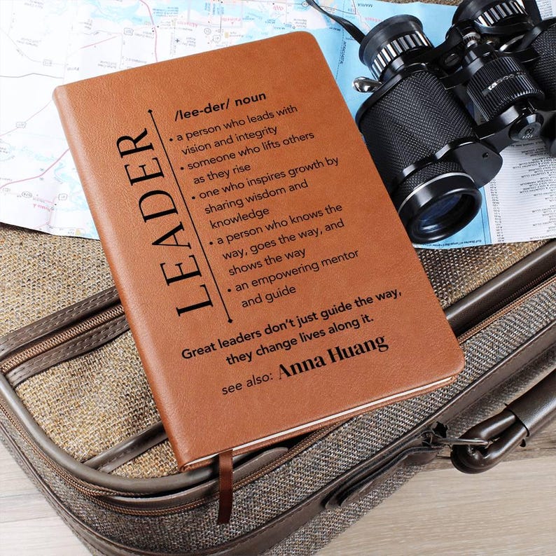 May include: A brown leather-bound journal with the word "LEADER" printed vertically. The journal includes a definition of "leader" and a quote. Binoculars and a map are visible in the background.