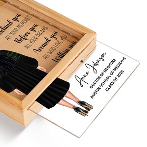 K&ouml;nnte beinhalten: Eine Geschenkbox zum Abschluss mit einer Illustration einer schwarzen und gr&uuml;nen Abschluss-Kappe und -Robe. Die Box hat einen transparenten Acryleinsatz mit dem Text "Anna Johnson, Doktor der Medizin, Austin School of Medicine, Klasse von 2025".