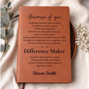 Puede incluir: Un cuaderno con cubierta de cuero color canela y texto impreso en negro que dice: Because of you, Challenges become opportunities, Confidence begins to grow, People feel supported, And lives are changed, Difference Maker, A person whose actions inspire, Steven Smith.