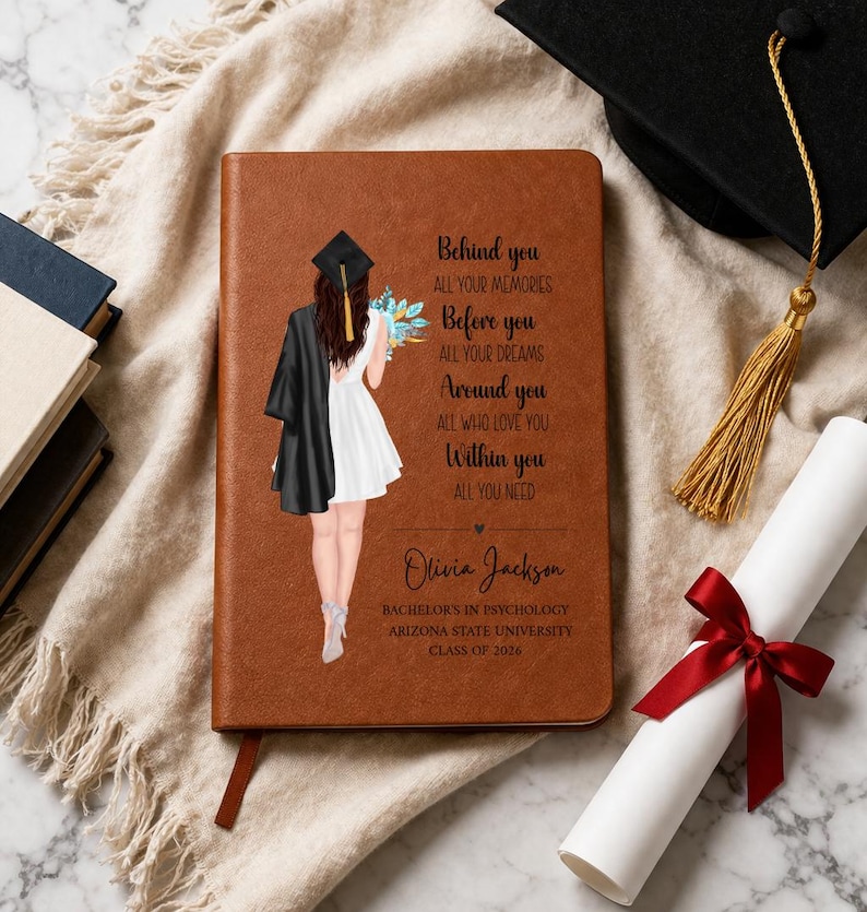 May include: A brown leather-bound journal with a graduation theme. The cover features an illustration of a graduate and text. A graduation cap, tassel, and diploma with a red ribbon are also visible.