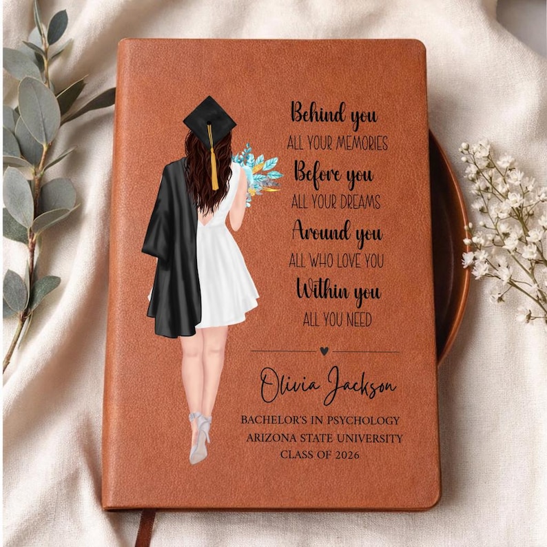 May include: A brown leather-bound journal with a graduation theme. The cover features an illustration of a graduate in a cap and gown, holding flowers. Text includes the graduate's name, degree, university, and class year.