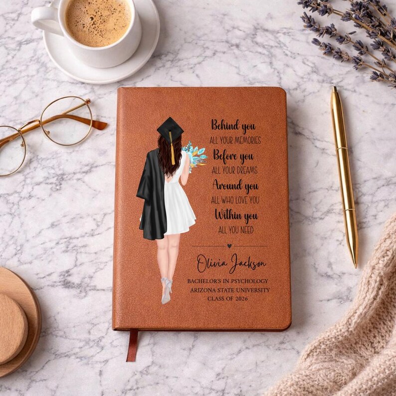 May include: A brown leather-bound journal with a graduation-themed illustration and text. The illustration depicts a graduate in a cap and gown. The text reads "Behind you, Before you, Around you, Within you." Includes the name "Olivia Jackson" and "Class of 2026."