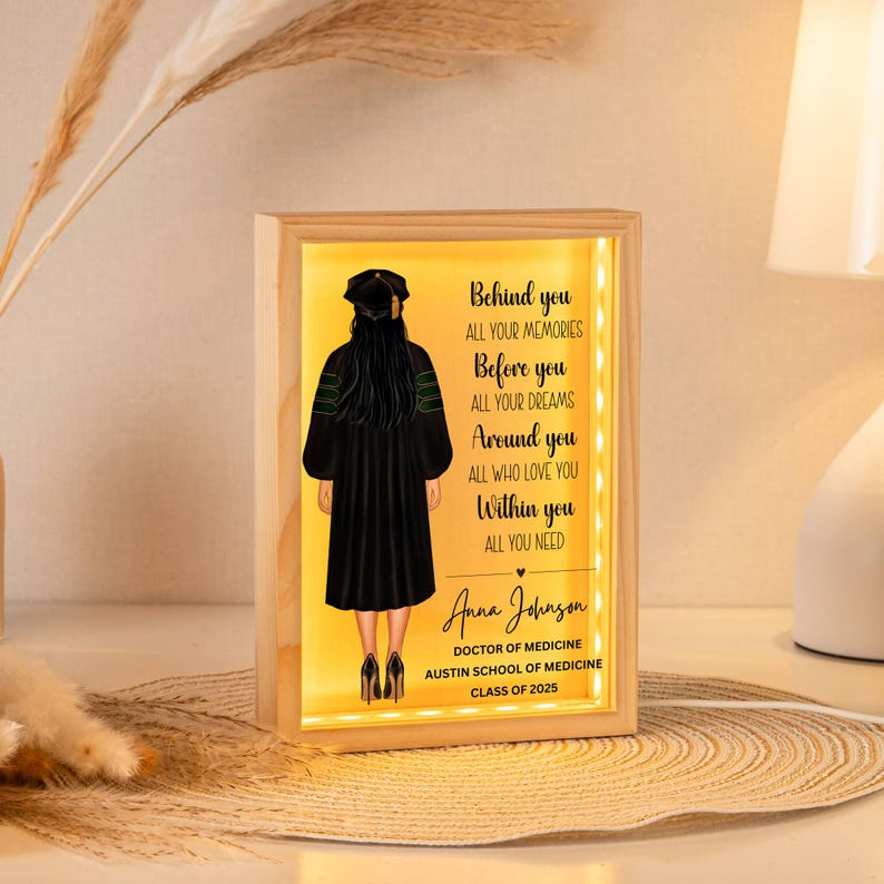 K&ouml;nnte beinhalten: Eine leuchtende Holzkiste mit einer Silhouette einer Abschluss-Kappe und dem Text "Behind you all your memories, Before you all your dreams, Around you all who love you, Within you all you need". Die Box enth&auml;lt auch den Namen "Anna Johnson", den Text "Doctor of Medicine, Austin School of Medicine, Class of 2025" und ein kleines Herz.