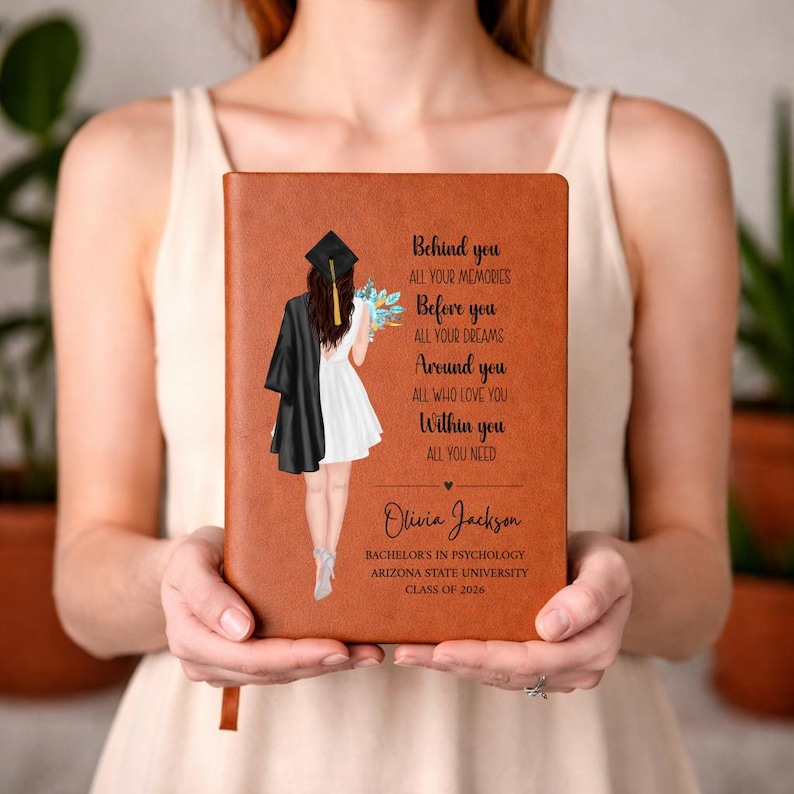May include: A brown leather-bound journal with a graduation-themed illustration and text. The illustration depicts a graduate in a cap and gown. The text reads "Behind you, all your memories. Before you, all your dreams." Includes a name, degree, university, and class year.