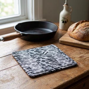 May include: A square, quilted hot pad with a black and white animal print pattern, including zebra stripes and leopard spots. The pad is on a wooden table, near a cast iron skillet and a loaf of bread.